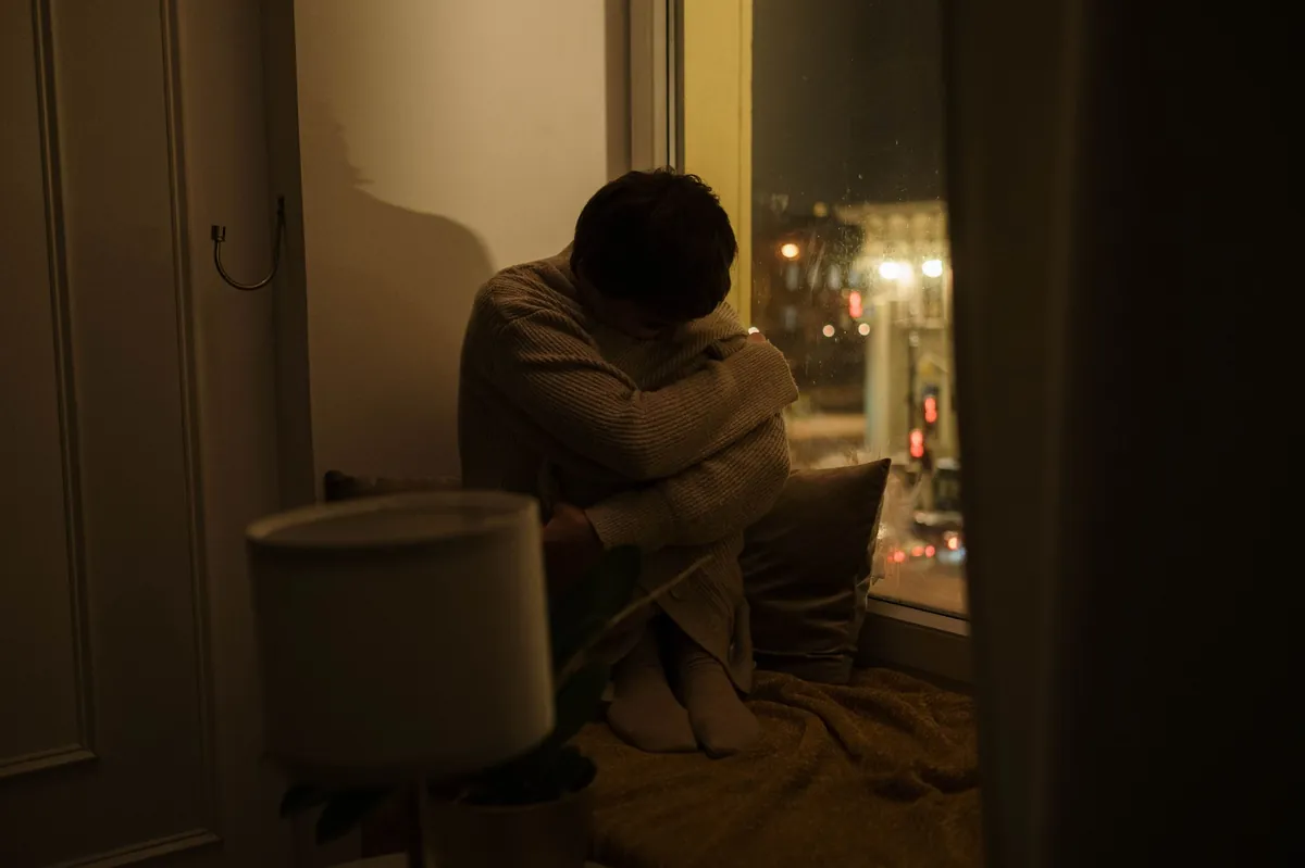 A person sitting alone at night beside a window, hugging their knees