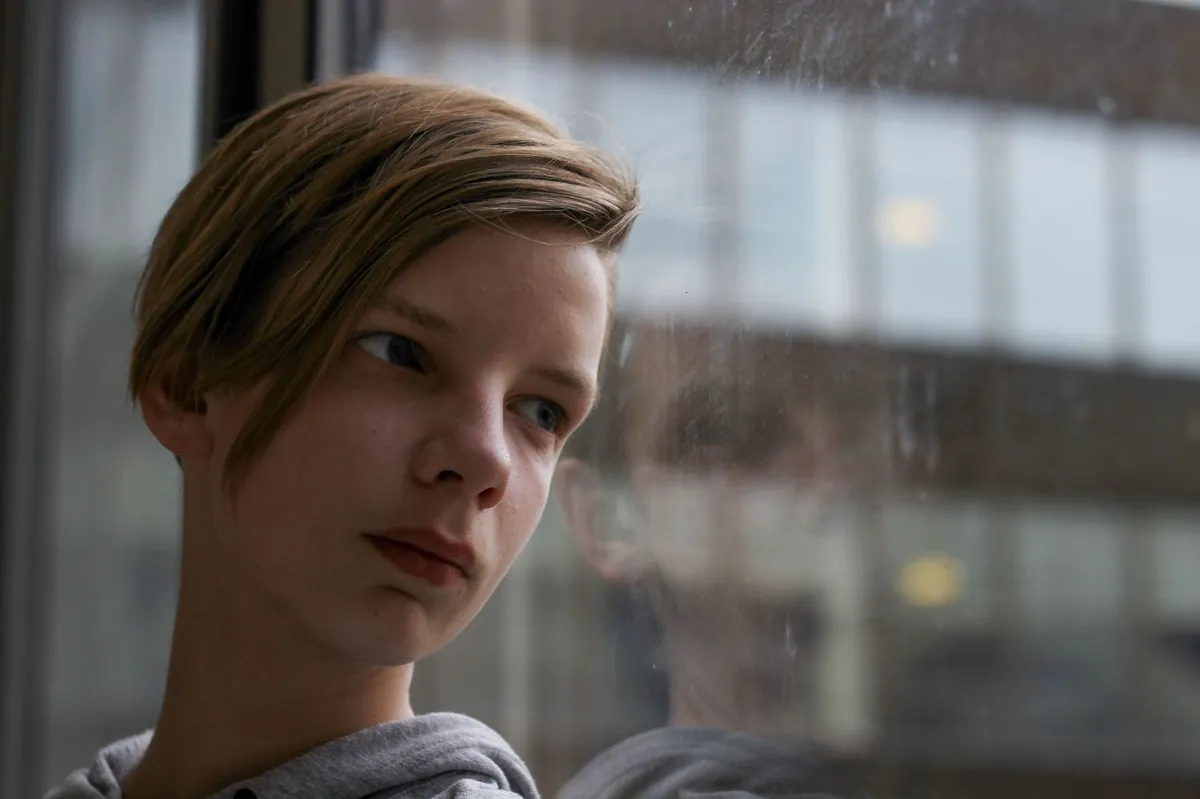 A teenager gazing through a rain-speckled window with a distant expression