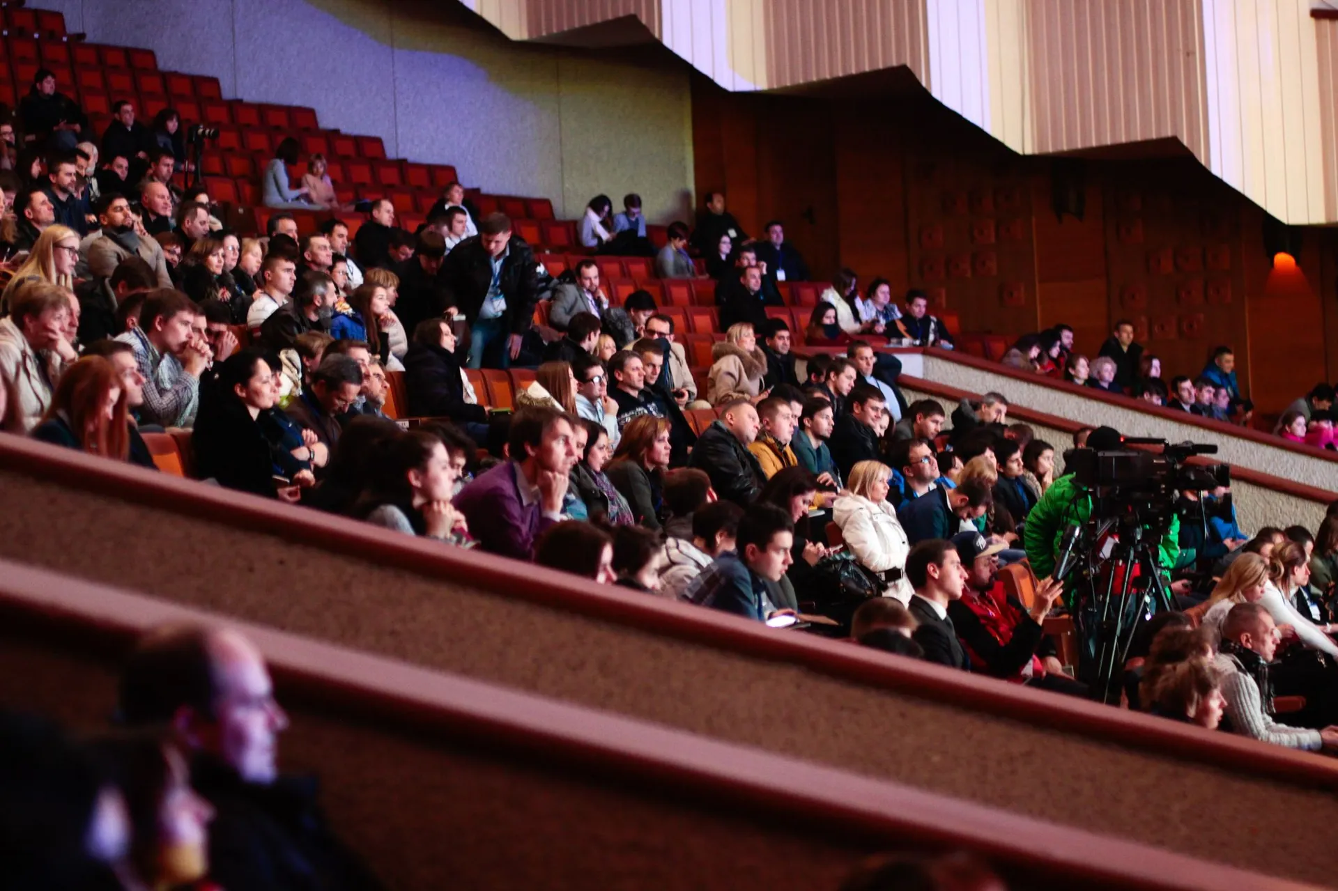 A full theater audience seen from the side, listening in warm light
