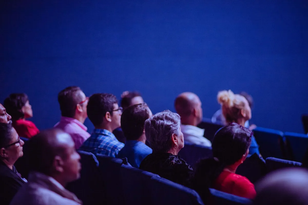 Rows of listeners in a dim auditorium lit by blue light